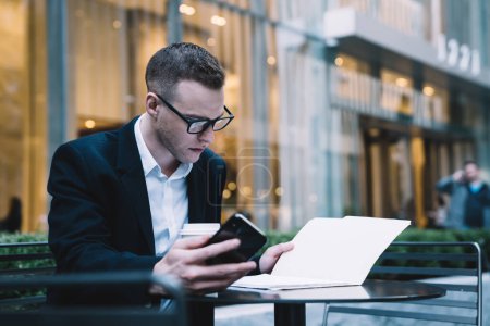 Joven empleado masculino concentrado en gafas y chaqueta de traje sentado a la mesa en la cafetería de la acera y leyendo documentos mientras sostiene el teléfono inteligente