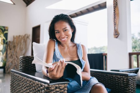 Photo for Asian happy intelligent satisfied female reading interesting book while resting in armchair at home and enjoying hobby looking at camera - Royalty Free Image
