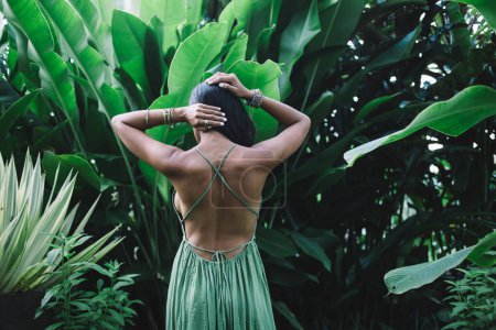 Rückenansicht einer unkenntlich sitzenden Brünetten mit nackten Schultern im grünen Kleid stehend und anrührenden Haaren vor dichten grünen tropischen Pflanzen
