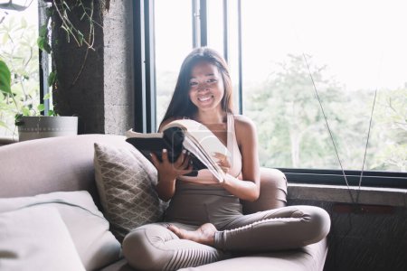 Photo for Young smart ethnic woman with crossed legs sitting on couch turning pages of book at home and smiling at camera - Royalty Free Image