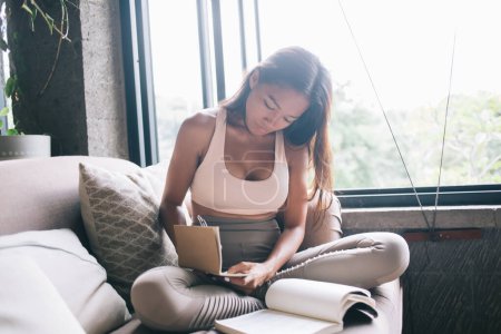 Photo for Young smart Asian lady with long dark hair making notes in notepad and reading book sitting at living room on couch with pillows - Royalty Free Image