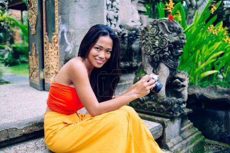 Foto de Joven asiática sonriente coqueta fotógrafa vestida con elegante traje de verano boho naranja sentada cerca de la estatua oriental y mirando a la cámara - Imagen libre de derechos