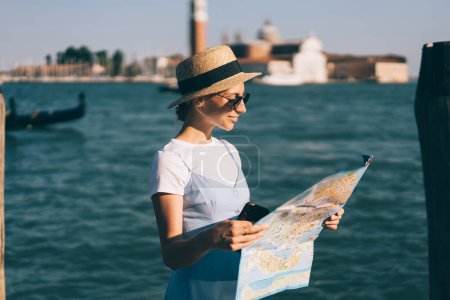 Youth Caucasian traveller in trendy sunglasses enjoying solo vacations for exploring romantic Italian city during recreation summer holidays, carefree woman 20s in hat reading navigation map