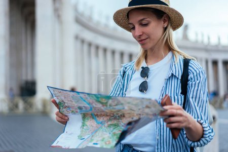 Attractive Caucasian tourist checking information from paper map for navigate own location during city sightseeing in ancient Rome,beautiful female traveller exploring Vatican city during Italian trip