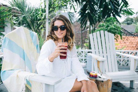 Gorgeous smiling positive woman in white dress and sunglasses drinking red summer beverage through straw while relaxing on chair in resort cafe
