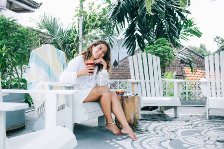 Smiling content joyful relaxed female leaning on hand while resting on chair in summer in recreation zone with red beverage
