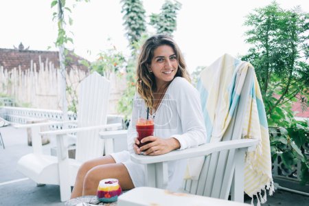 Smiling positive content young female tourist chilling on terrace cafe with glass of red smoothie and color dessert looking at camera