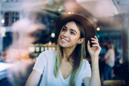 Foto de Señora atractiva y alegre en ropa casual sentado en la cafetería y sonriendo mientras toca el sombrero y mirando a través del vidrio - Imagen libre de derechos
