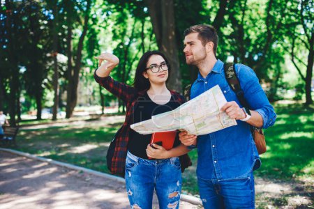 Photo for Young woman in casual wear and spectacles raising arm showing direction to handsome male traveler, couple in love spend vacation on journey together holding map for getting to city destinations - Royalty Free Image