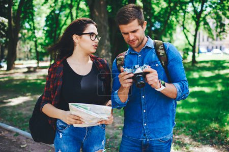 Photo for Young woman in casual wear talking to her boyfriend checking pictures on camera having tour in city on weekends, couple in love resting visiting city using map and taking photos while exploring - Royalty Free Image