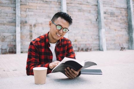 Photo for Happy young Asian man in glasses and checkered shirt looking through book while standing and enjoying free time in street - Royalty Free Image