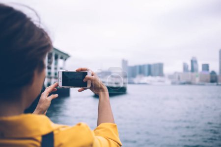 Foto de Vista trasera de una turista morena usando una blusa amarilla fotografiando ferry y rascacielos en la pintoresca bahía de la ciudad de Hong Kong en un teléfono celular blanco en un día desagradable - Imagen libre de derechos