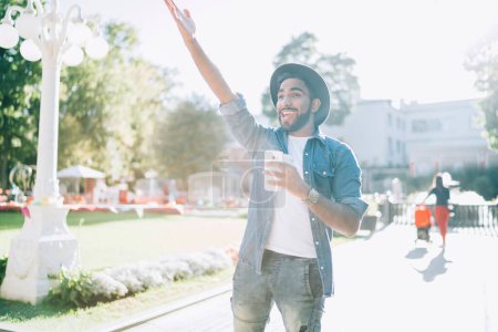 From below excited stylish Hispanic guy in denim clothes with takeaway hot drink and smartphone smiling away and greeting friend while standing on street against blurred view summer park