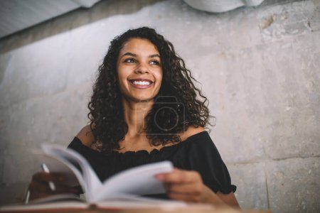 From below of happy ethnic female in casual clothes flipping through pages of notepad sitting at table in cafe looking away