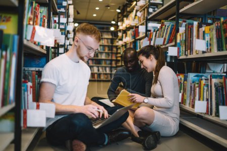 Grupo multirracial de jóvenes universitarios positivos que estudian en el suelo entre librerías en la biblioteca con ordenador portátil y libros de texto preparándose para las lecciones