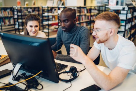 Téléchargez les photos : Jeunes impliqués intelligents étudiants diversifiés naviguant sur l'ordinateur à la table de la bibliothèque pour créer un rapport éducatif conjoint partager des idées et des connaissances avec enthousiasme - en image libre de droit