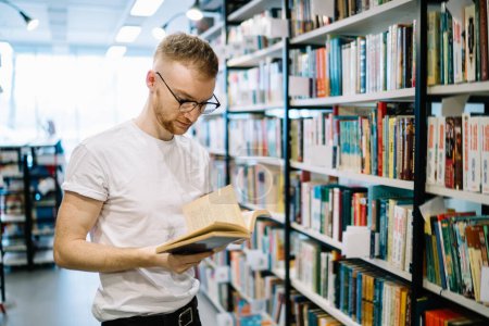 Téléchargez les photos : Homme barbu concentré réfléchi calme en chemise blanche et lunettes tenant et lisant livre tout en se tenant debout dans la bibliothèque moderne lumière - en image libre de droit