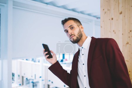 Bearded male manager in red suit and with smartphone leaning over railing and looking at camera while working in spacious office