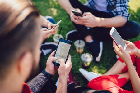 Group of young friends texting on mobile phones sitting around on grass with cups of coffee in city in summer