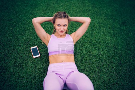 Téléchargez les photos : Femme optimiste musclée confiante en vêtements de sport légers avec smartphone souriant et faisant des craquements abdominaux sur le champ d'herbe et regardant la caméra - en image libre de droit