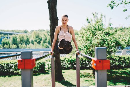 Young blond sportive female raising legs on parallel bars for training abdominal muscles in park at summertime and looking confidently in camera