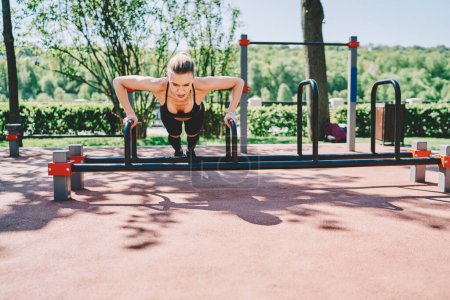 Confident female in sport clothes doing push up exercise on parallel bar doing fitness at sports ground in park on sunny day