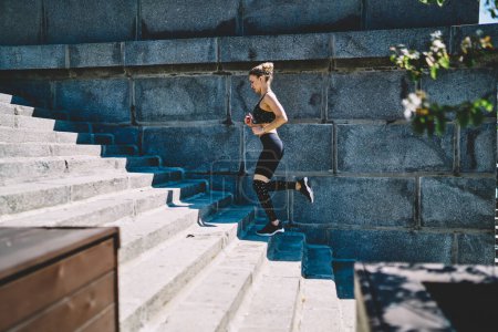 Téléchargez les photos : Vue latérale d'une athlète féminine concentrée en survêtement noir à la mode qui court sur des escaliers à bascule au stade extérieur lors d'une chaude journée d'été - en image libre de droit