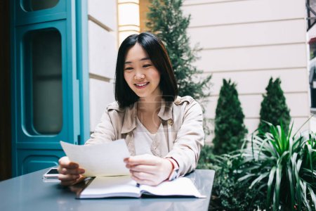 Foto de Joven mujer asiática alegre con el pelo corto negro sonriendo y sentado en el escritorio con cuaderno mientras lee el documento en la calle de la ciudad - Imagen libre de derechos