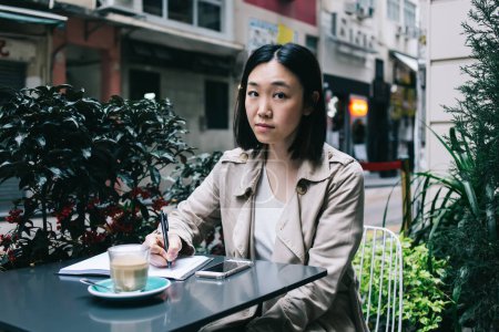 Foto de Mujer asiática joven de pelo corto en ropa casual sentada en el escritorio con taza de café y teléfono inteligente mientras escribe en el portátil y mirando a la cámara en la calle de la ciudad - Imagen libre de derechos
