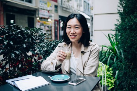 Photo for Calm glad ethnic Asian woman in jacket resting at table with smartphone and copybook while sipping coffee on terrace in restaurant and looking at camera on blurred background - Royalty Free Image