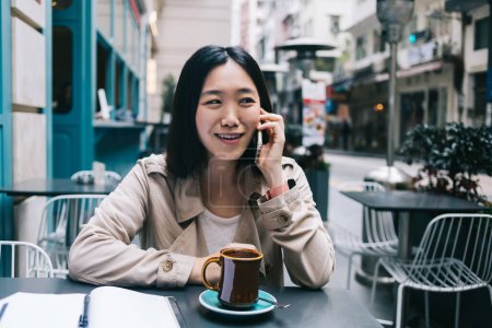 Foto de Alegre mujer asiática en ropa casual hablando en el teléfono inteligente mientras está sentado en la mesa con una taza de café en la cafetería mirando hacia otro lado - Imagen libre de derechos