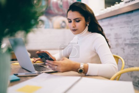 Konzentrierte ernsthafte brünette Frau in lässiger weißer Kleidung überprüft Informationen mit Handy und Laptop, während sie tagsüber Hausaufgaben in der Cafeteria macht