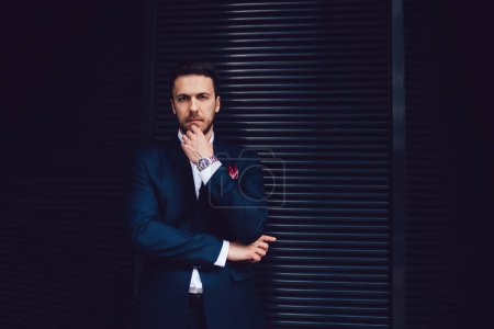 Pensive serious executive bearded businessman in suit looking at camera and making decision while touching face with hand and standing at wall