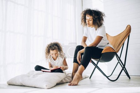 African American mother and daughter looking down on journal and writing while sitting on floor pillow and soft chair in light room