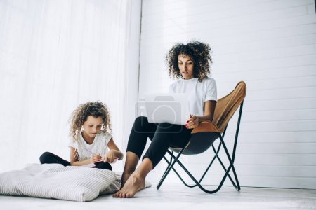 Curly ethnic daughter sitting on floor pillow and writing in journal while mother sitting in soft armchair near and using laptop