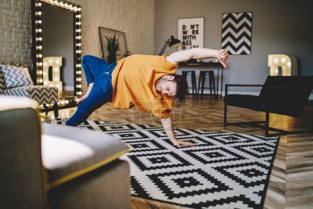 Foto de Hombre activo en ropa casual practicando postura de yoga en alfombra haciendo deporte apoyado en la mano en casa moderna mirando hacia abajo - Imagen libre de derechos