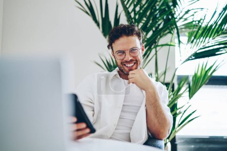 Sonriente barbudo hombre de anteojos usando el teléfono móvil mientras está sentado en la mesa delante de la computadora portátil poniendo la mano en la barbilla y mirando a la cámara