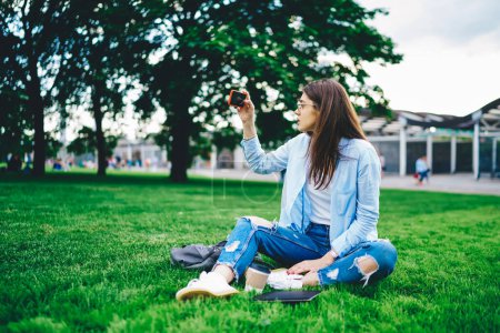 Foto de Mujer joven en ropa casual usando cámara de teléfono inteligente filmando video para blog sentado en el parque verde en tiempo libre, mujer influencer tomando selfie en el teléfono móvil comunicándose con los seguidores en vacaciones - Imagen libre de derechos