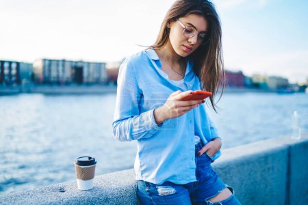 Foto de Turista femenina pensativa en ropa casual enviando mensajes de texto a través de un teléfono inteligente que descansa en el puente del río, joven blogger hipster revisando el correo y enviando mensajes de texto en el teléfono móvil al aire libre - Imagen libre de derechos