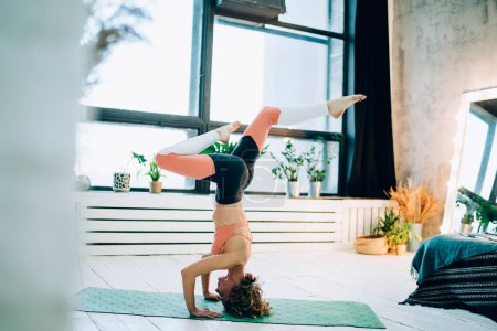 Strong female in active wear standing in pose for training balanse during workout in home apartment, sportswoman doing headstand exercise for practising static dressed in trendy tracksuit