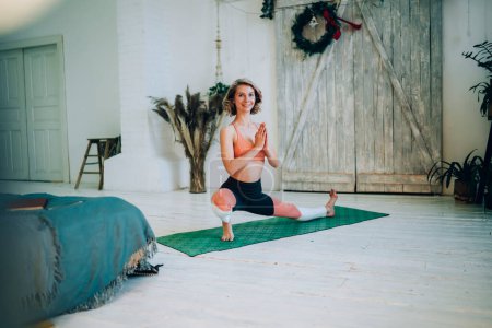 Happy woman in trendy sports clothing enjoying time for training stretching skills standing in pose on mat in loft home apartment, smiling female with positive thinking doing flexibility workout