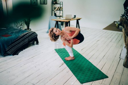 Von oben starke schlanke, konzentrierte Frau in Sportbekleidung, die Stuhl-Yoga-Pose auf Matte macht, mit gefalteten Händen zu Hause nach oben schauend