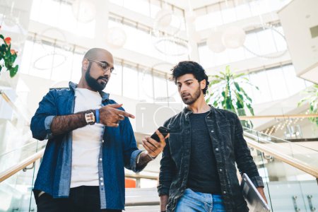 Ethniker demonstriert neues Smartphone und erklärt Freund mit Laptop Vorteile, während er im modernen Büro auf verschwommenem Hintergrund die Treppe hinuntergeht