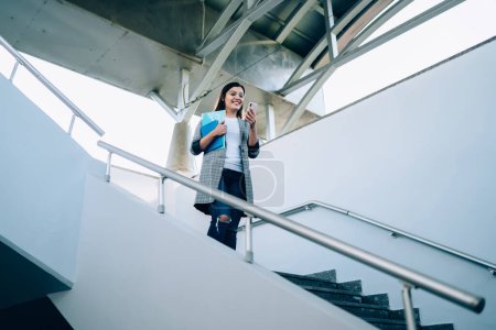 Smiling caucasian young woman reading notification on mobile phone walking down on urban staircase, cheerful trendy dressed millennial hipster girl happy about getting good 4G internet send text message