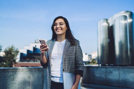 Portrait of cheerful caucasian hipster girl in trendy wear satisfied with 4G connection chatting online during trip, smiling woman laughing at funny content from social networks using cellphone