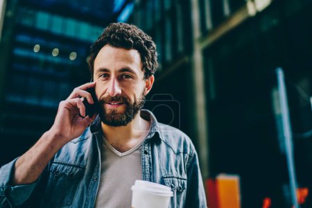 Foto de Bajo ángulo de contenido barbudo empresario masculino en ropa casual mirando a la cámara mientras habla en el teléfono móvil en la calle de la ciudad moderna - Imagen libre de derechos