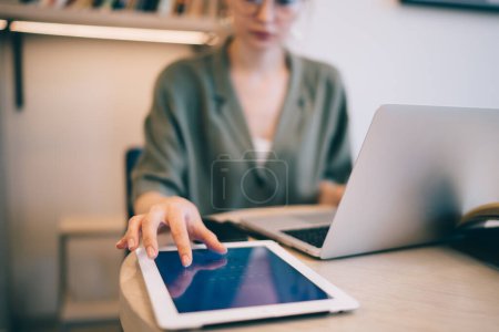 Foto de Mujer de la cosecha sentada en la mesa tocando la pantalla de la tableta mientras usa el ordenador portátil en el espacio de trabajo - Imagen libre de derechos