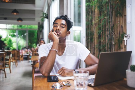 Foto de Estudiante étnico cansado sentado en la cafetería en la mesa con teléfono inteligente portátil y pedazo de papel y pensando mientras sostiene la cabeza con la mano y mirando a la cámara - Imagen libre de derechos