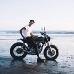 Side view of Hispanic young calm cheerful modern guy relaxing on black motorcycle on sunset ocean beach looking at camera