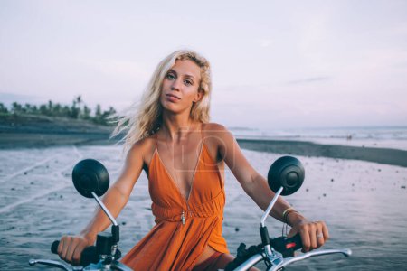 Photo for Attractive seductive young female with long hair in orange dress sitting on motorbike and looking at camera on seashore at dusk - Royalty Free Image